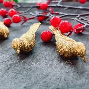 May include: Four gold bird figurines on a gray surface with red berries and branches in the background.