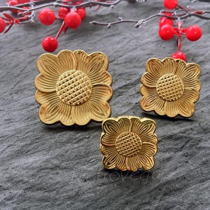 May include: Three gold-colored metal flower-shaped decorative embellishments. The embellishments are square and have a raised, textured center.