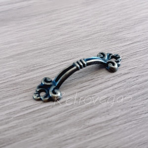 May include: A small, ornate, antique-style, metal drawer pull with a curved design and decorative details. The pull is finished in a bronze color.