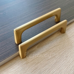 May include: Two gold-toned cabinet pulls with a rectangular shape and rounded edges. The pulls are made of metal and have a brushed finish.