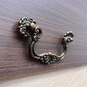 Peut inclure: Gros plan d'une poignée de tiroir en bronze antique avec des détails floraux ornés. La poignée incurvée est fixée à une surface en bois foncé. Le mot "Retrovegas" est imprimé sur la surface en bois.