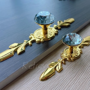 May include: Two gold cabinet pulls, each featuring a light blue crystal knob. The pulls have an ornate, leaf-like design. The pulls are on a grey surface. The word "Retrovi" is visible in the image.