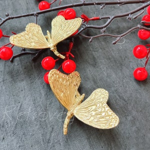 Peut inclure: Deux broches en forme de papillon dorées avec des détails complexes. Les broches sont sur un fond de tissu gris foncé avec des baies rouges.