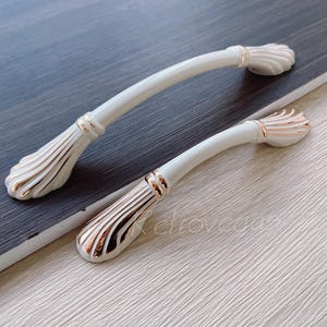 May include: Two white ceramic cabinet pulls with gold accents. The pulls have a decorative, scalloped design.