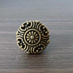 以下が含まれることがあります： 装飾的な花柄のデザインが施された、丸いアンティーク真鍮色のキャビネットノブ。ノブは、渦巻く模様に囲まれた中央の花を特徴としています。「Retrovegas」の文字が表面に筆記体で書かれています。