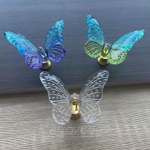 May include: Three decorative butterfly cabinet knobs. Two knobs have blue and green wings, and one is clear. Each butterfly has a gold-coloured base. The knobs are made of a translucent material.