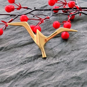 Puede incluir: Una escultura de pájaro de metal dorado en forma de grúa. La escultura está sentada sobre una superficie gris texturizada con bayas rojas y ramas en el fondo.