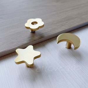 May include: Three gold-toned metal cabinet knobs with different shapes: a star, a flower, and a crescent moon.