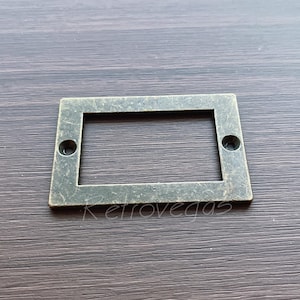 May include: A rectangular metal frame with a worn, antique finish. The frame has a rectangular opening in the centre, perfect for displaying a small label or tag.