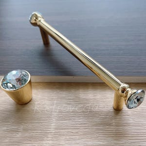 May include: Gold cabinet hardware with a crystal knob. The handle is a long, thin, gold bar with a crystal on each end.