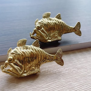 May include: Two gold-coloured fish-shaped drawer pulls with a detailed, textured surface. The fish have a wide mouth and large eyes.