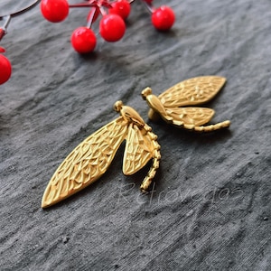 May include: Two gold-toned dragonfly earrings with intricate details. The earrings are on a grey textured surface.