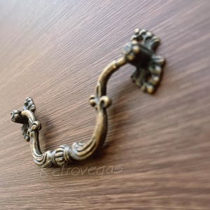 May include: A decorative antique-style cabinet pull with a ornate design. The pull is made of metal and has a brass finish.
