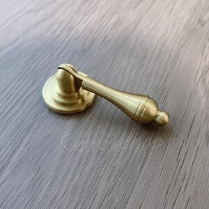 May include: A gold-coloured cabinet pull with a round base and a decorative handle. The hardware is made of metal and has a classic design. The pull is shown on a light grey wood surface.