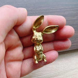 Può includere: Una statuetta di coniglio dorata con un viso sorridente e le braccia incrociate. Il coniglio è in piedi su una superficie piana.