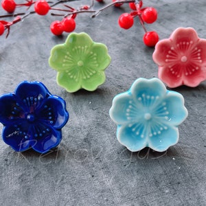 May include: Four ceramic flower-shaped chopstick rests in blue, green, pink, and light blue. The flower-shaped rests are on a gray surface, with red berries and a branch in the background. These are ideal for Asian-themed table settings.