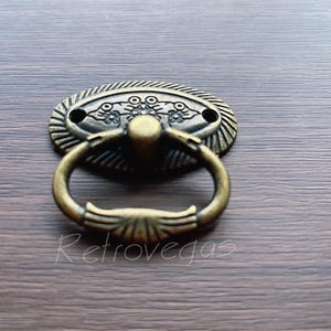 May include: Antique brass drawer pull with an oval-shaped base and a ring handle. The base has a decorative floral design.