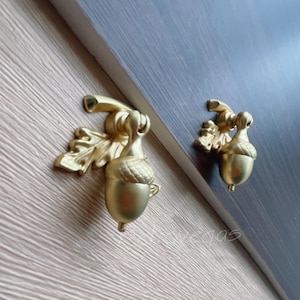 Op de afbeelding: Twee gouden kastknoppen in de vorm van een eikel met een bladontwerp. De knoppen zijn gemonteerd op een donkergrijs oppervlak.