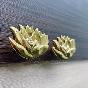 Może przedstawiać: Dwa złote gałki do szafek w kształcie kwiatu lotosu. Gałki wykonane są z metalu i mają szczegółowy, trójwymiarowy wzór. Gałki zamontowane są na ciemnobrązowej drewnianej powierzchni.