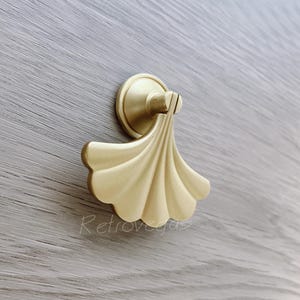 May include: A gold-toned cabinet pull with a fan-shaped design. The pull is mounted on a round base.