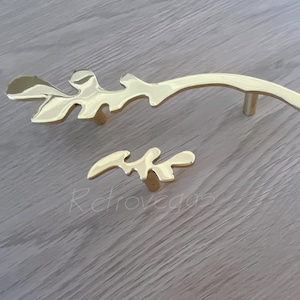 May include: Two gold-toned metal cabinet pulls in the shape of stylised leaves. The larger pull is curved and has a small, round knob on the bottom. The smaller pull is straight and has a more detailed leaf design.