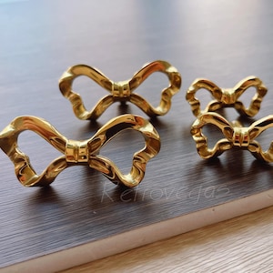 Op de afbeelding: Vier goudkleurige, strikvormige decoratieve objecten. De strikken hebben een getextureerde, metallic afwerking en zijn gerangschikt op een donker oppervlak met houtnerf. De strikken zijn van verschillende grootte.