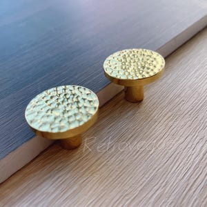 Op de afbeelding: Twee gouden kastknoppen met een gehamerde afwerking. De knoppen zijn rond en hebben een verhoogde rand. Ze worden getoond op een houten oppervlak.