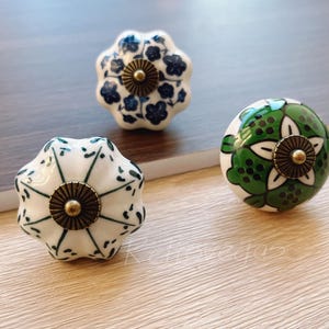 Puede incluir: Tres pomos de cerámica con diseños florales. Los pomos son blancos con patrones florales azules y verdes. Cada pomo tiene un acento de latón en el centro.