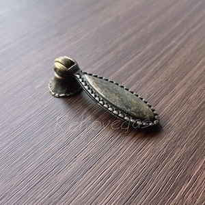 Può includere: Una piccola maniglia per armadio di forma ovale in stile antico con un bordo decorato con perline. La maniglia è in metallo con una finitura bronzo scuro e un accento dorato nella parte superiore.