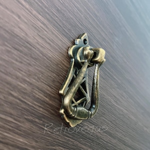 Può includere: Maniglia per cassetto color ottone antico con un design decorativo. La maniglia presenta un'impugnatura curva e una placca posteriore ornata, montata su una superficie in legno scuro. La parola "Retrovegas" è visibile.