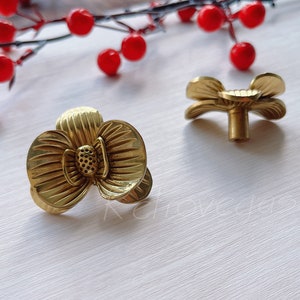 May include: Two gold-toned flower-shaped cabinet knobs with a detailed design. The knobs are on a white surface.