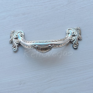 May include: Silver drawer pull with a decorative, ornate design. The pull is shaped like a half-circle with a heart-shaped centre.