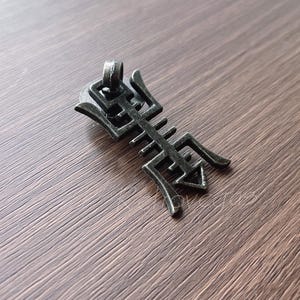 May include: A dark grey metal pendant with an intricate, stylised design. The pendant has a loop at the top for a chain and features a central bar with symmetrical, angular extensions. The pendant is displayed on a wooden surface.