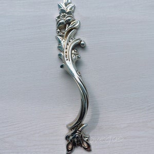 May include: A silver-coloured, ornate cabinet handle with a floral and leaf design. The handle curves gracefully, with detailed embellishments along its length. The handle is likely made of metal and is set against a light-coloured background.
