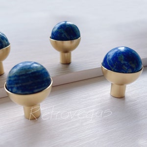 Op de afbeelding: Vier gouden kastknoppen met blauwe lapis lazuli stenen toppen. De knoppen zijn rond en hebben een gladde, gepolijste afwerking.