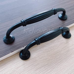 May include: Two black metal cabinet door pulls with a curved design. The pulls have a fluted texture and are approximately 4 inches long.