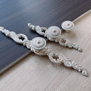 Op de afbeelding: Drie sierlijke witte en gouden kastknoppen met een decoratief bloemmotief. De knoppen hebben een ronde vorm en een verhoogd midden.