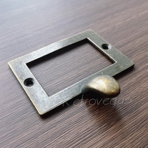 以下が含まれることがあります： アンティークブラスの引き出しプル、長方形のラベルホルダーと丸いハンドル付き。