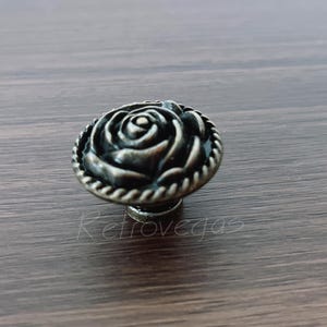 Puede incluir: Pomo de armario de estilo antiguo con diseño de rosa. El pomo es de metal con un acabado oscuro y envejecido. La rosa está detallada con múltiples pétalos y un borde en forma de cuerda. El pomo está montado sobre una superficie de madera.