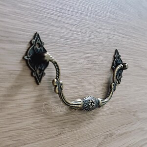 May include: A decorative, ornate, antique-style drawer pull with a black finish and a curved design. The pull is made of metal and has a diamond-shaped plate on one end.