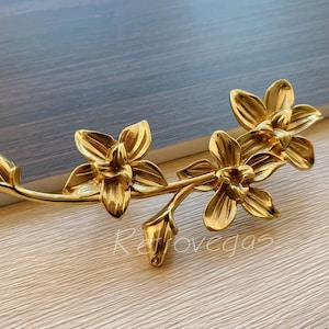Könnte beinhalten: Goldfarbener Metall-Schubladengriff mit floralem Design. Der Griff verfügt über drei stilisierte Blüten mit jeweils fünf Blütenblättern.