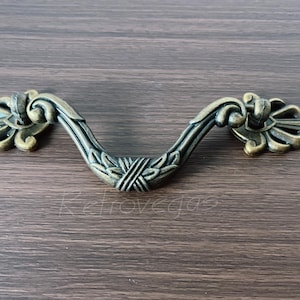 May include: An antique brass-effect drawer pull with decorative floral detailing. The curved handle has a central design and is set against a dark wood grain background. A vintage-style hardware piece.