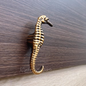 May include: A bronze seahorse-shaped cabinet pull. The seahorse is detailed with vertical ridges and a curved tail. The pull is mounted on a dark brown wooden surface. The seahorse is approximately 10 cm tall.