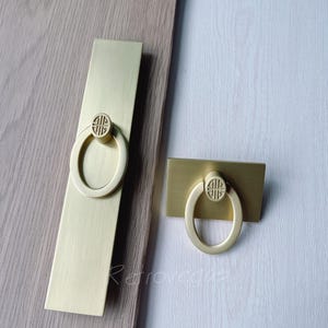 May include: Two gold-toned cabinet pulls with a ring design. The larger pull is rectangular and the smaller pull is square.