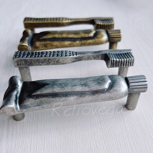 May include: Three metal drawer pulls shaped like a tube of toothpaste. The pulls are in different finishes: antique brass, silver, and aged silver.