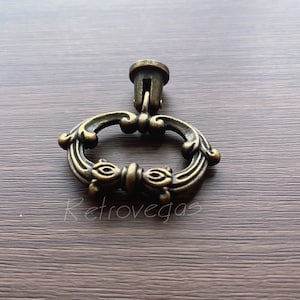 Peut inclure: Une poignée de tiroir décorative en métal doré ornée d'une anse en forme d'anneau. La poignée présente un motif floral et est fixée à une petite base ronde.