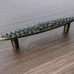 May include: An antique bronze cabinet pull with a woven texture design. The pull has two posts for mounting and is approximately 5 inches long. The Retrovegas logo is visible in the lower right corner.