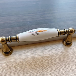 May include: A white ceramic cabinet knob with a floral design and gold accents. The knob is rectangular in shape and is attached to a gold metal base.