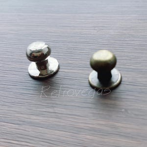 Pode incluir: Dois botões de armário, um prateado e outro dourado, com a parte superior redonda e a base plana.