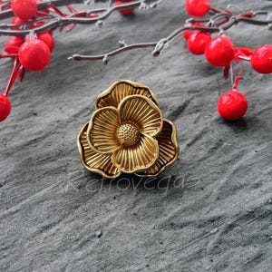 Op de afbeelding: Een ring van goudkleurige metaal met een bloemmotief. De bloem heeft vijf bloemblaadjes en een centrum met een reliëfmotief. De ring ligt op een grijze achtergrond.